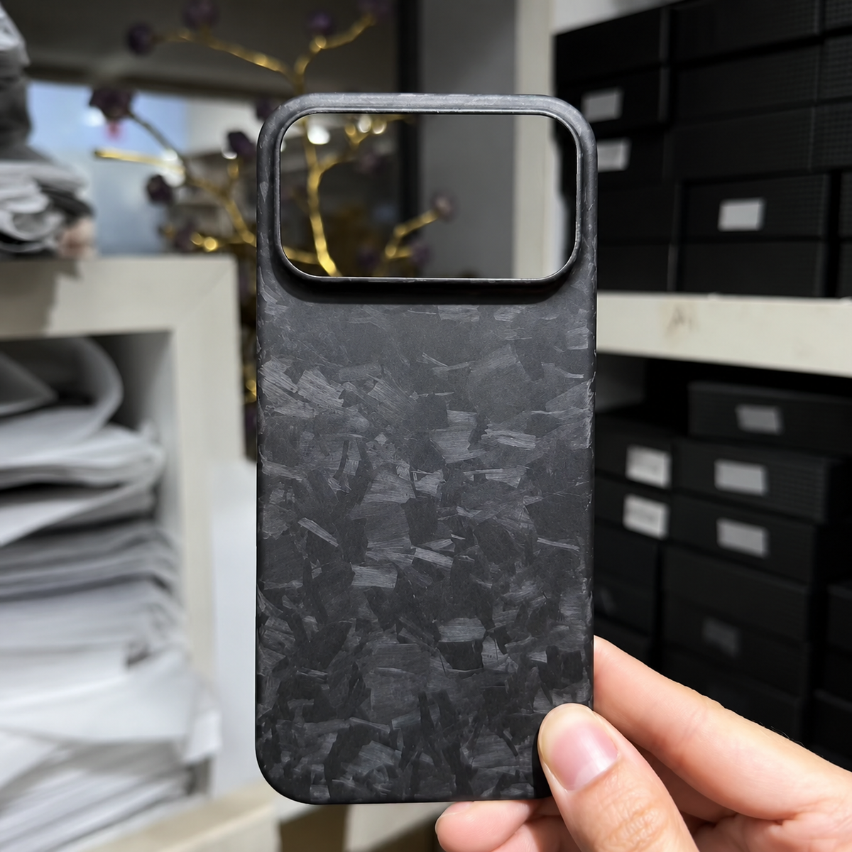 A hand holds a matte black phone case with irregular gray shard-like patches in front of shelves and stacked boxes. Its wide rectangular camera opening faces the viewer.