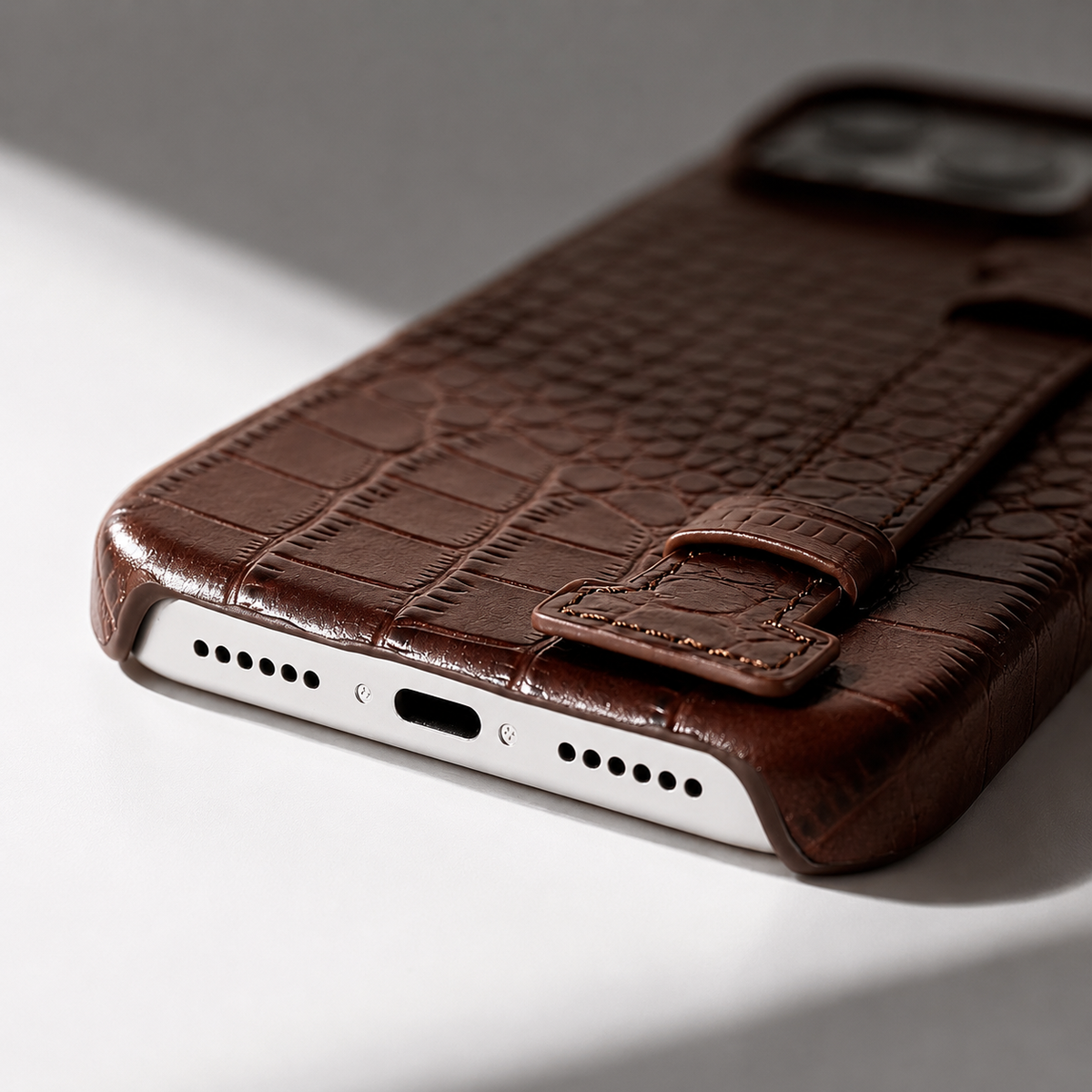A close-up of a brown crocodile-style case's lower edge shows stitching, the flat back strap and openings around the charging port and speakers.