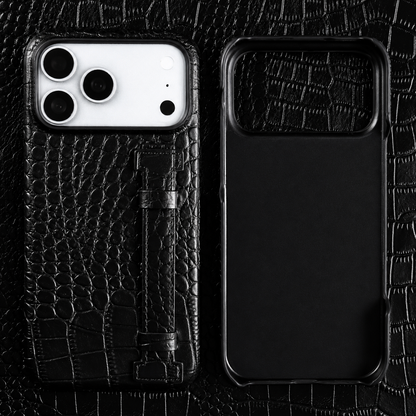 A black textured case and empty matching shell lie on black crocodile-patterned material. A vertical stitched strap sits along the case's back.