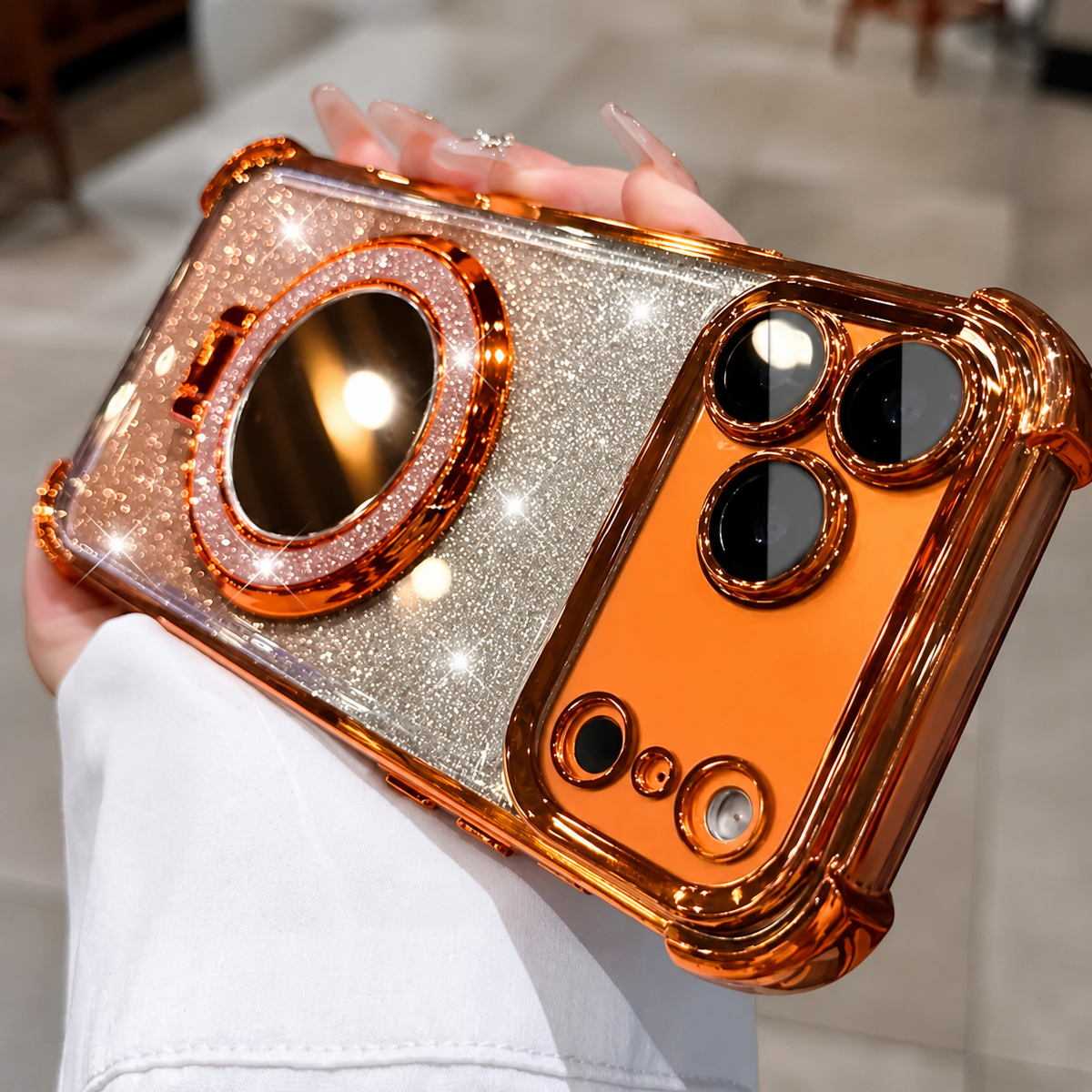 A hand holds a copper-orange phone case horizontally. Silver and champagne-colored glitter covers the back around a large round mirror, while orange trim frames the edges and camera openings.