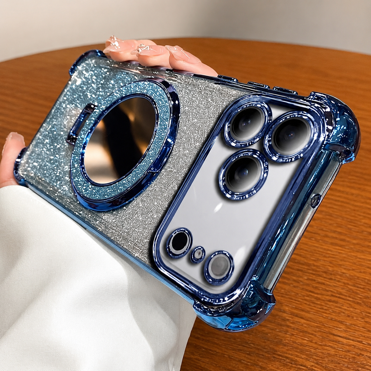 A hand holds a blue-trimmed silver glitter phone case horizontally in a café. The case has a large circular mirror on its back and matching blue trim around the camera panel.
