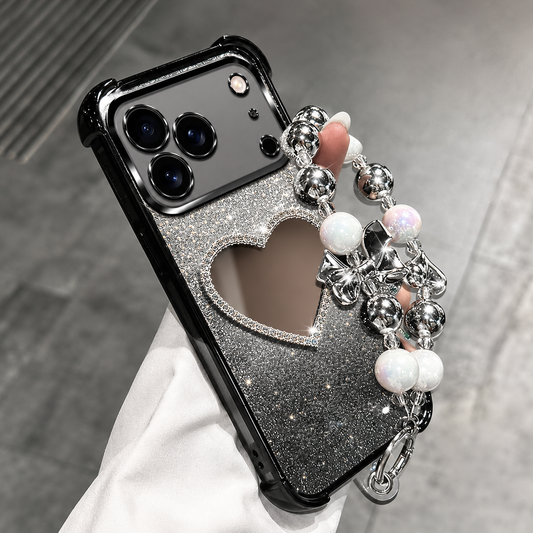 A hand holds a black glitter phone case over gray pavement. Its heart-shaped mirror has a sparkling border, and a beaded wrist loop combines shiny silver-colored beads, white pearl-like beads and bow charms.