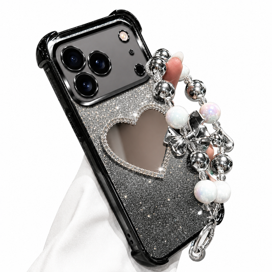 A hand in a white sleeve holds a black glitter phone case against white. A heart-shaped mirror sits beneath its glossy camera panel, and a wrist loop of silver-colored beads, pearl-like beads and bow charms hangs along the side.