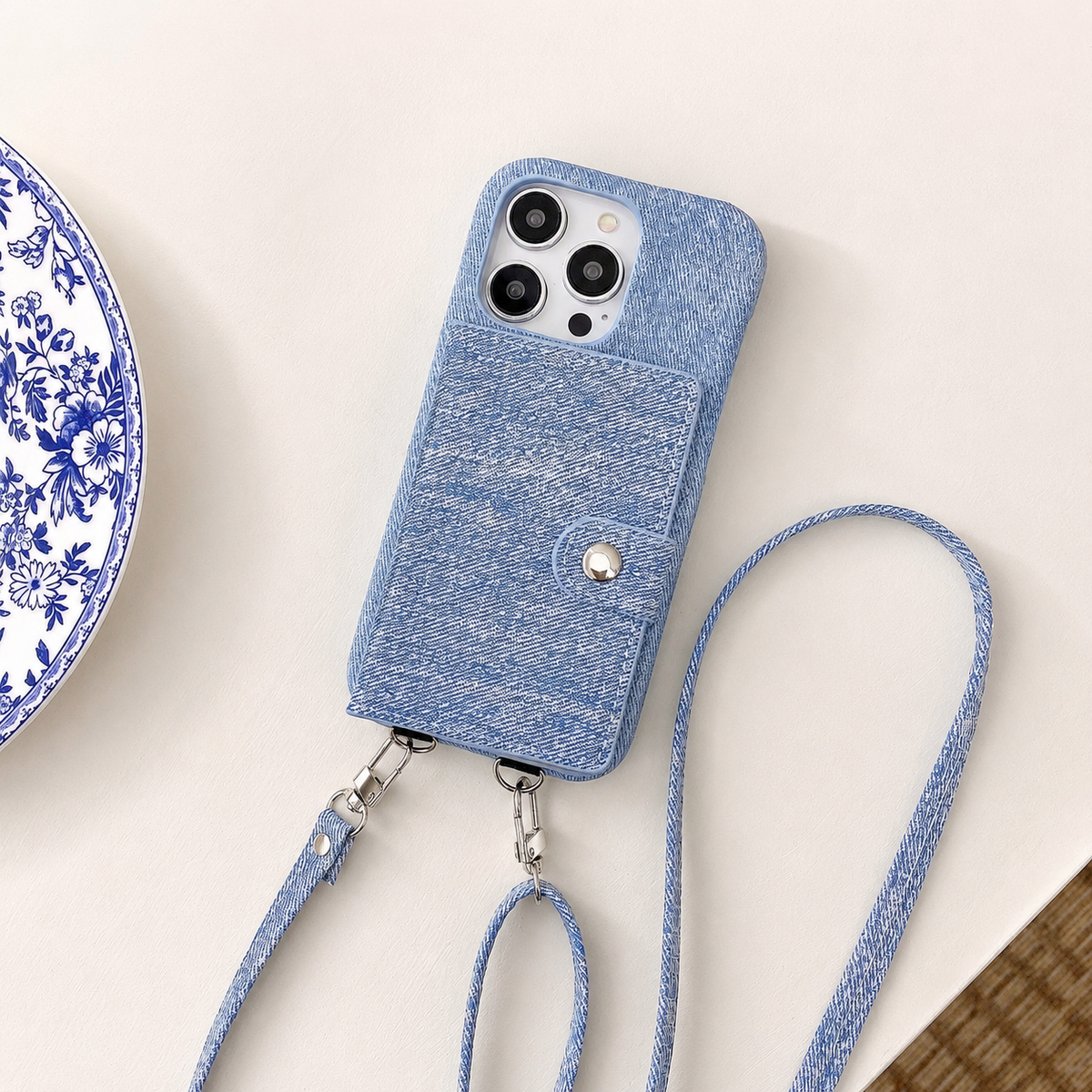 A blue woven-textured case lies rear-up on a pale table beside a patterned plate. A slim snap-tab wallet panel covers the back, and two detachable straps extend below.