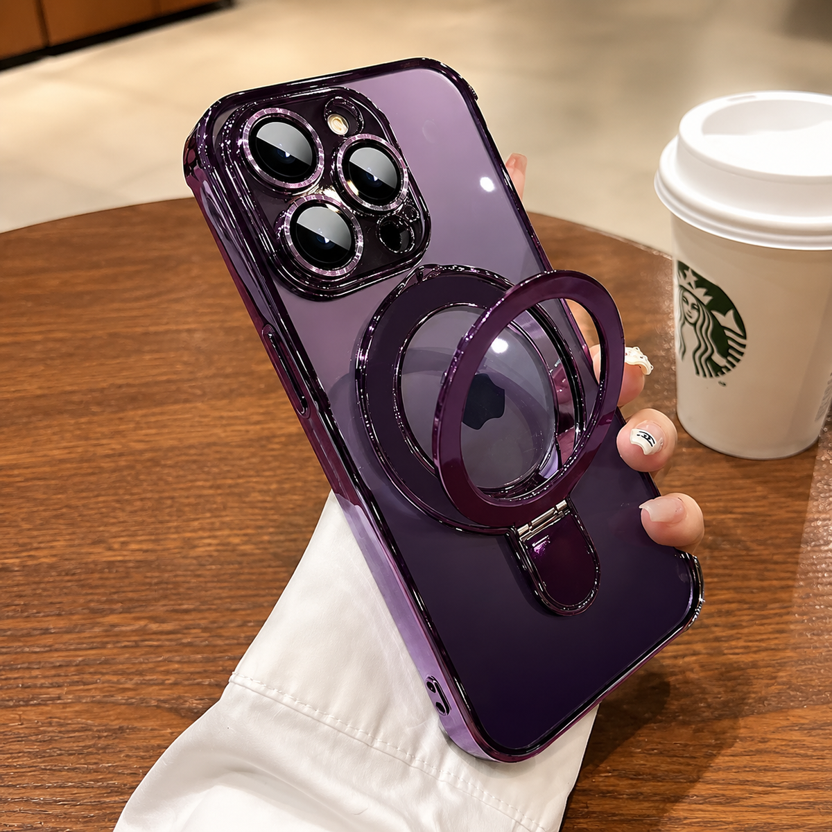 A hand holds a purple clear-cased phone above a wooden table, with its circular ring stand pulled outward into a loop.