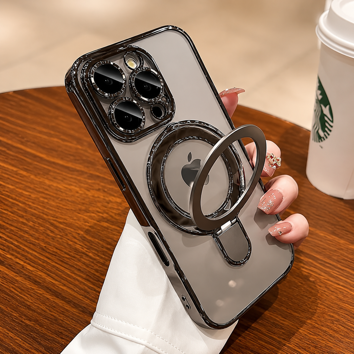 A hand holds a silver-gray clear-cased phone, showing its circular back ring unfolded. A takeaway cup sits on the cafe table behind it.