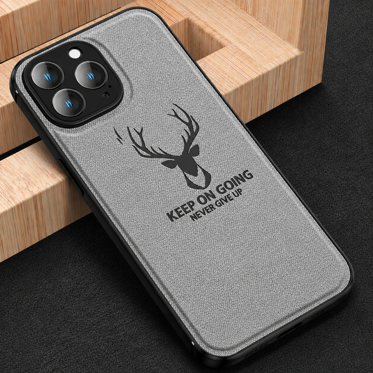 A gray fabric-textured phone case leans against a geometric wooden block on black fabric. A black stag head appears above 'Keep on going. Never give up,' beneath its three-lens camera surround.