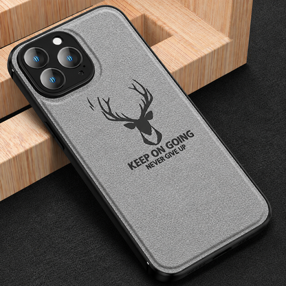 A gray fabric-textured phone case leans against a geometric wooden block on black fabric. A black stag head appears above 'Keep on going. Never give up,' beneath its three-lens camera surround.