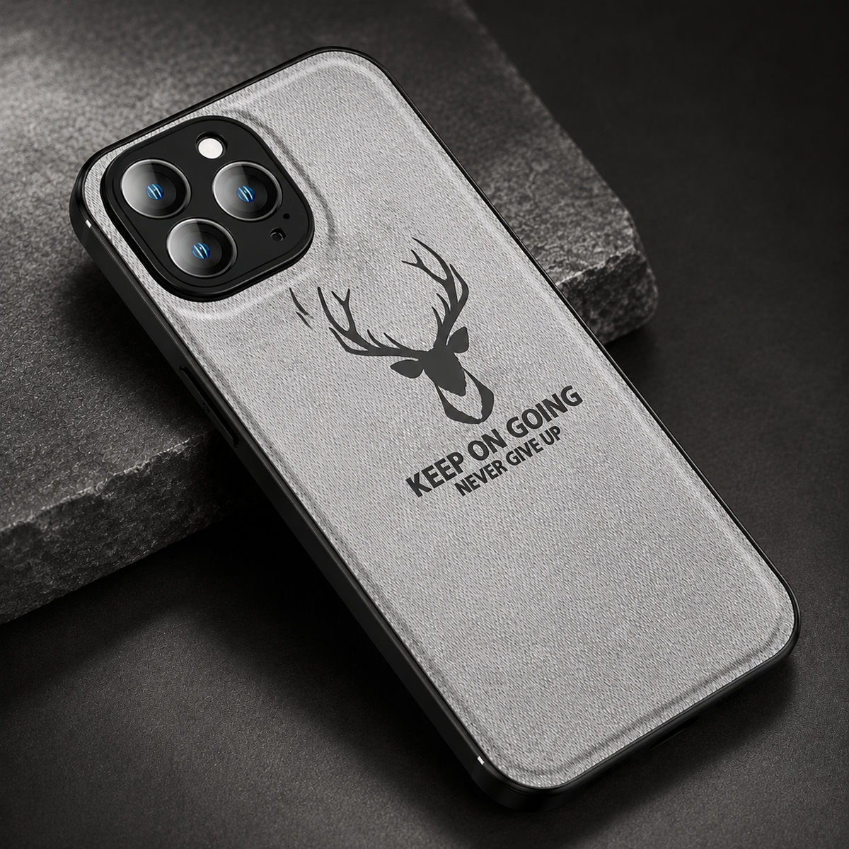 A gray fabric-textured phone case lies diagonally on dark stone. A black stag head appears above the words 'Keep on going. Never give up,' beneath a three-lens camera opening.