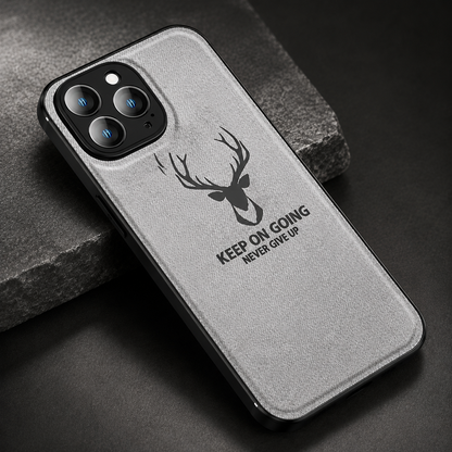 A gray fabric-textured phone case lies diagonally on dark stone. A black stag head appears above the words 'Keep on going. Never give up,' beneath a three-lens camera opening.