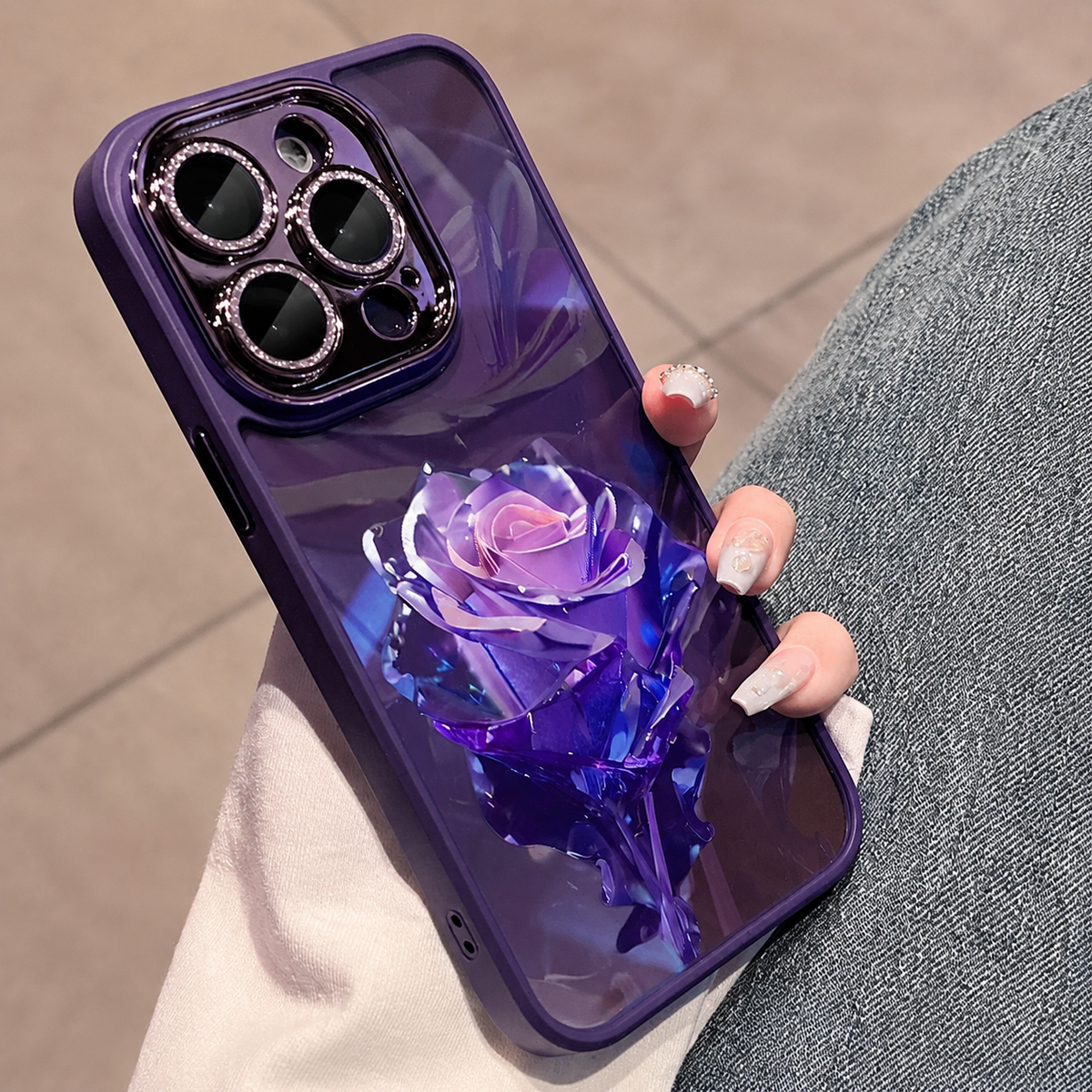 A hand holds a purple case beside blue jeans, decorated with a large purple faceted rose and matching leaves.
