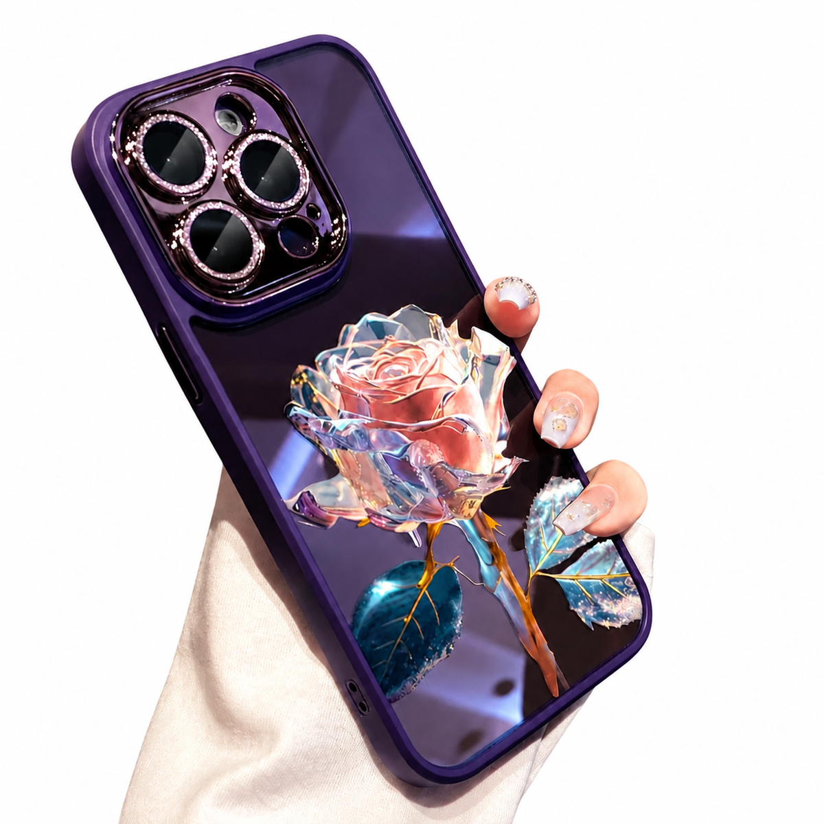 A hand holds a glossy purple case on white, decorated with a faceted pink-and-clear rose and blue-green leaves. Sparkling-looking rings outline the three camera lenses.