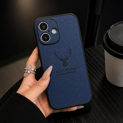 A hand with rings holds a navy textured case above a dark table beside a lidded cup. Its two-lens camera opening and deer emblem are visible.