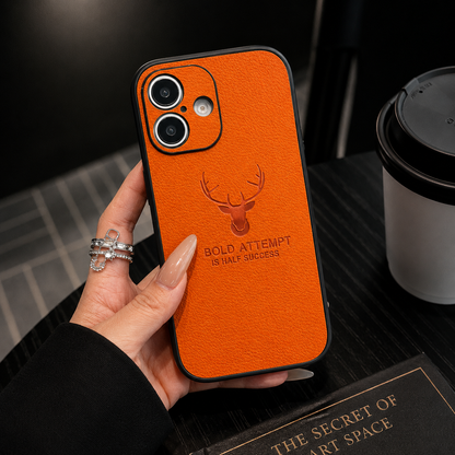 A hand holds an orange textured case above a dark table beside a lidded cup, showing a two-lens opening and embossed deer emblem.