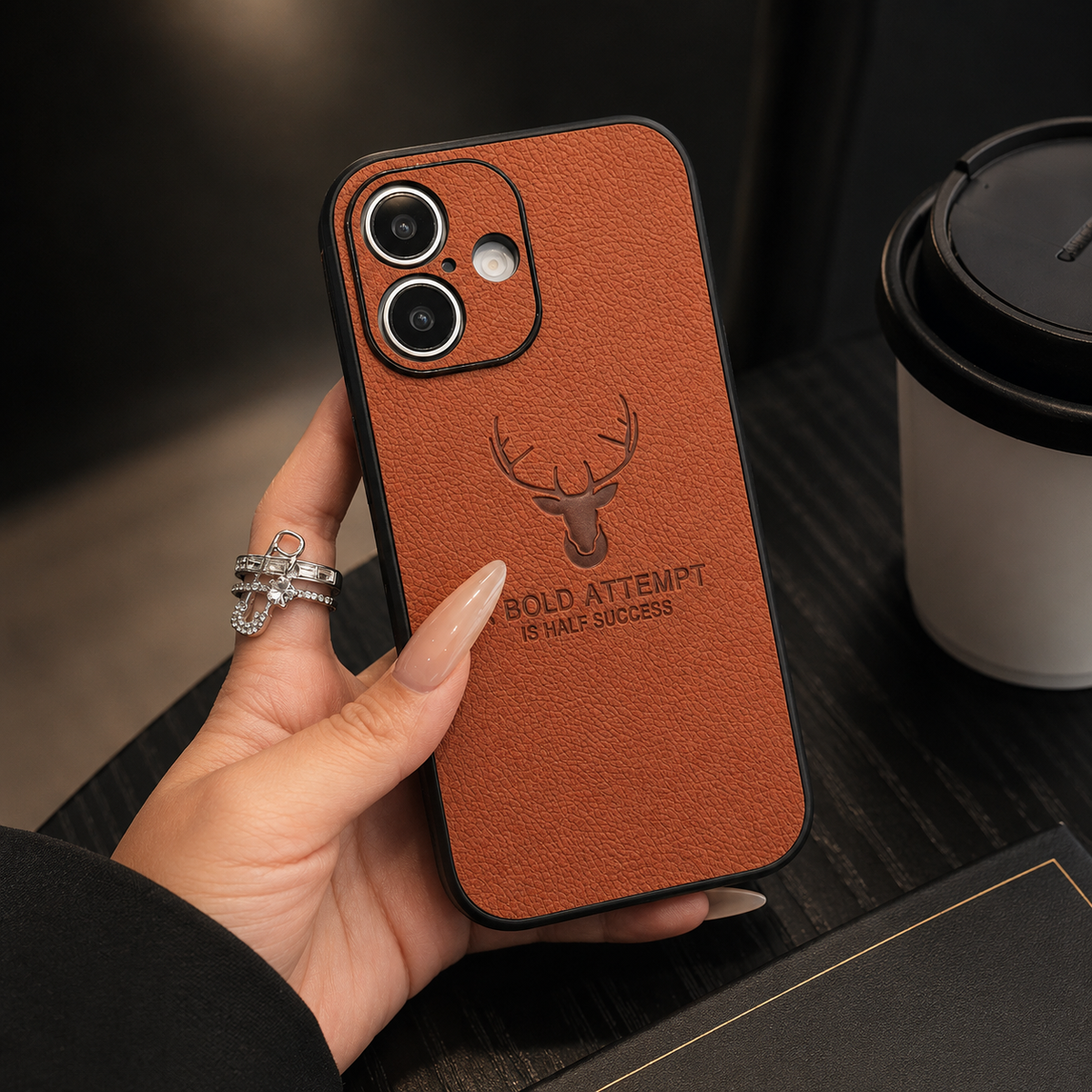 A hand holds a reddish-brown textured case above a dark table, showing its two-lens camera opening and deer emblem.