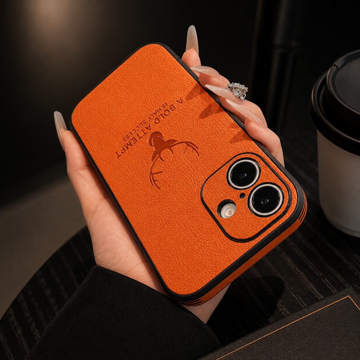 A hand holds an orange case horizontally, displaying its embossed deer emblem, short text and two-lens camera opening.