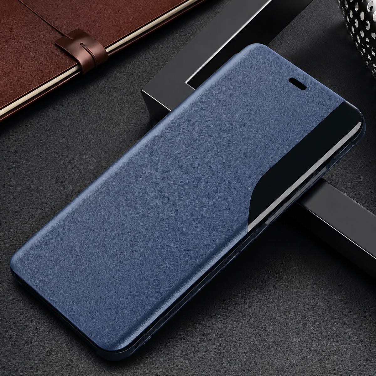 Blue flip phone case lying diagonally on dark blocks beside a notebook, with an earpiece slit and curved glossy black side strip.