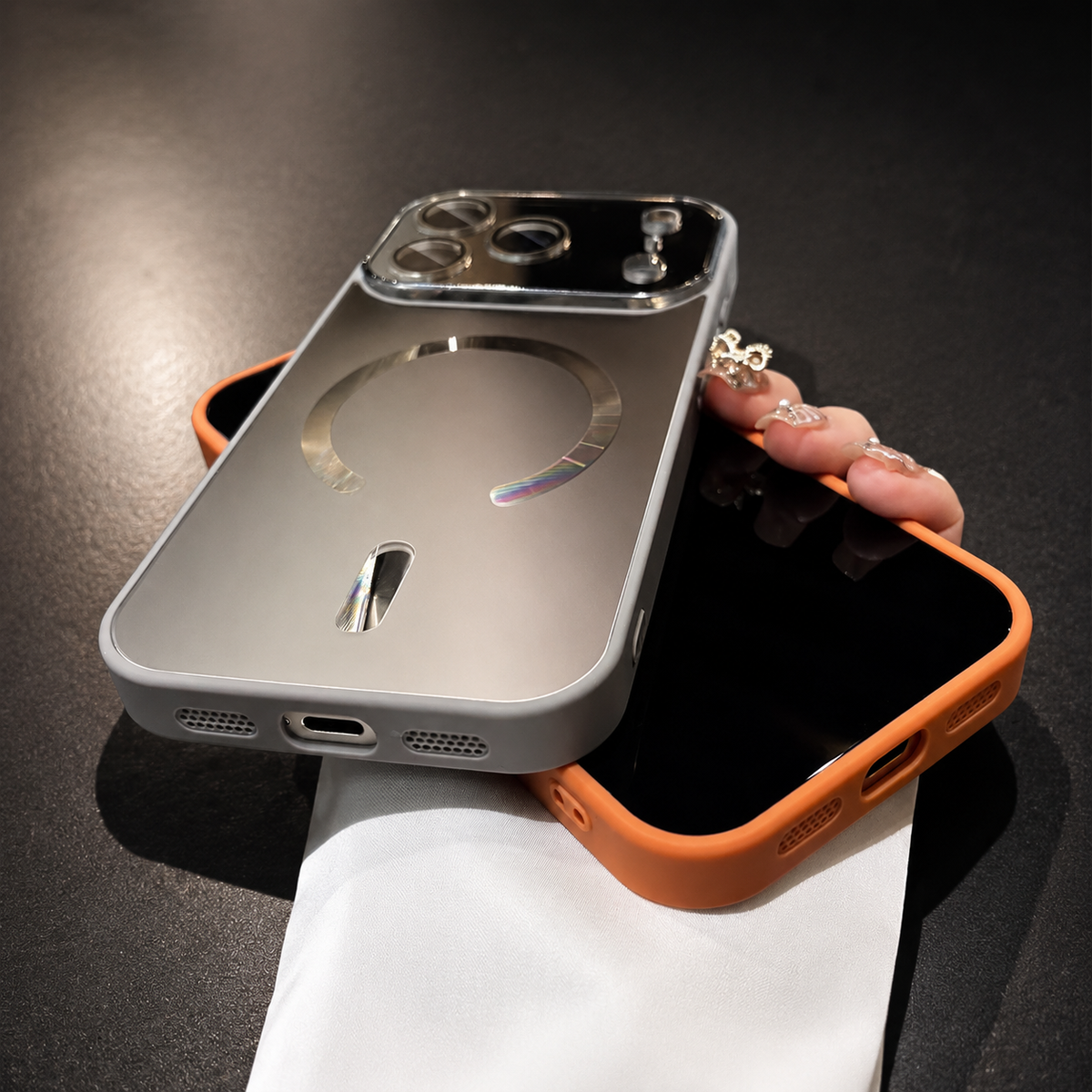 Silver-gray case rests rear-forward across an orange-cased phone in a hand above a dark tabletop. The lower edge shows charging and speaker openings.