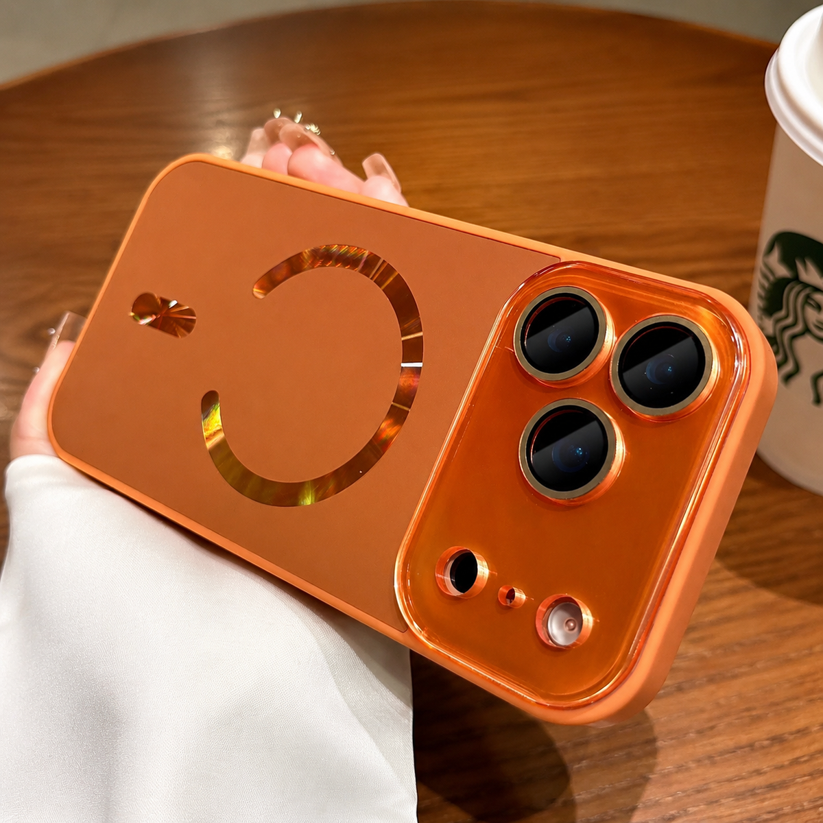 Hand holds an orange case horizontally above a wooden café table. A reflective magnetic ring and alignment bar decorate its back, and three lenses have raised surrounds.