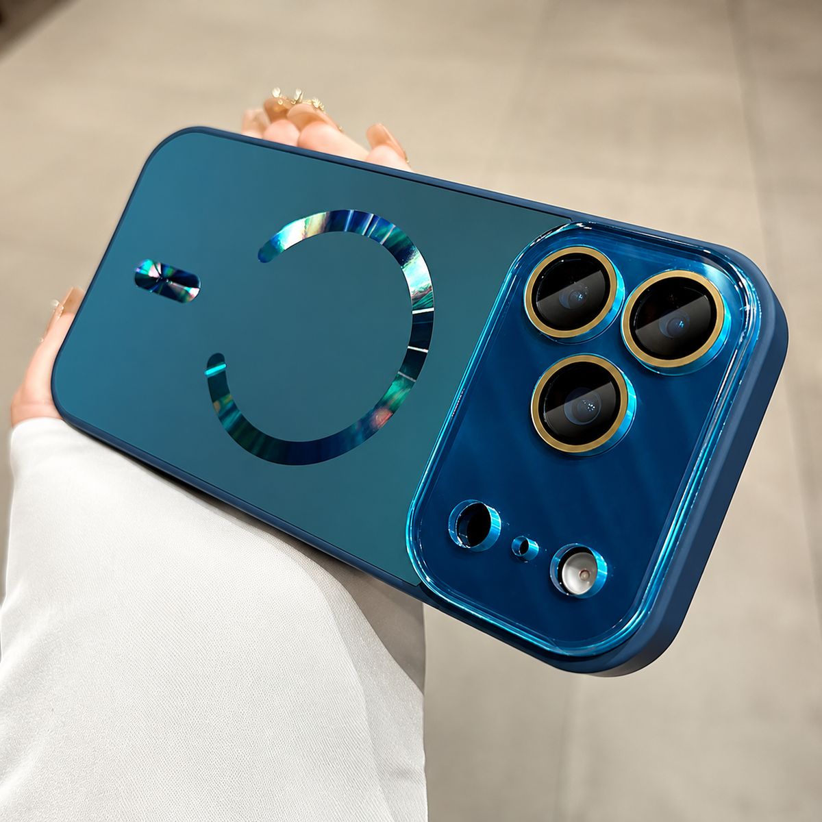 Hand holds a blue case horizontally indoors. A reflective blue magnetic ring and alignment bar decorate the back beside a broad glossy camera panel.