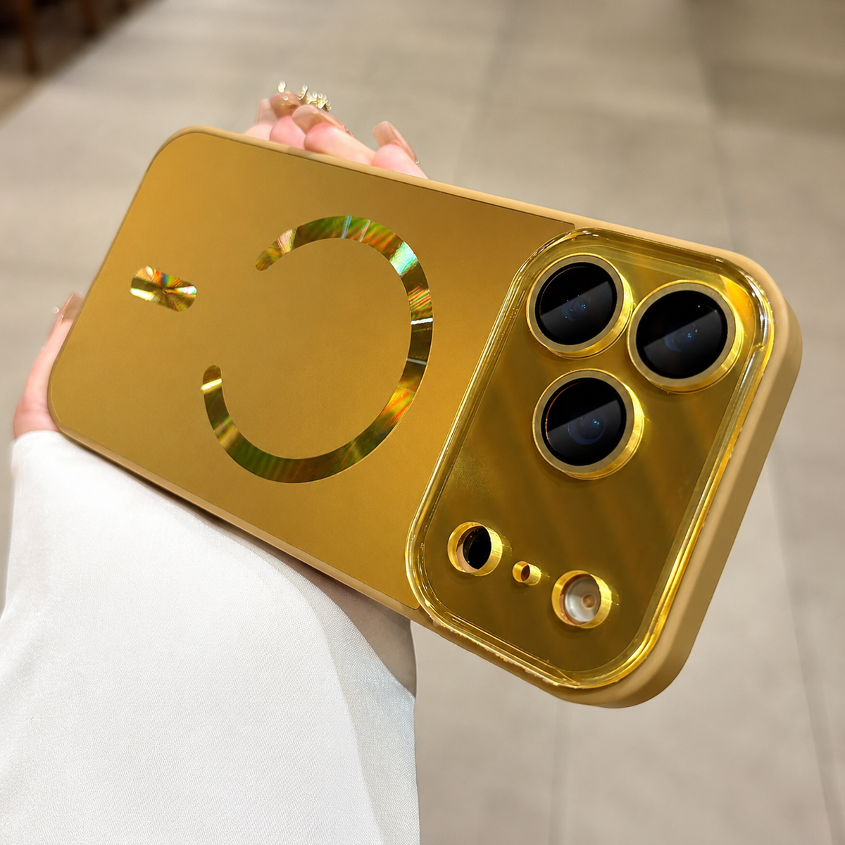 Hand holds a yellow case horizontally indoors. A reflective gold-colored magnetic ring and alignment bar sit beside its broad glossy camera panel.
