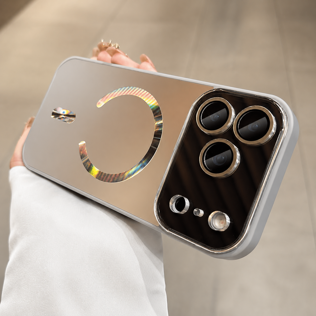 Hand holds a silver-gray case horizontally indoors. Its reflective magnetic ring and alignment bar catch rainbow colors beside a dark camera panel with gold-colored lens rings.