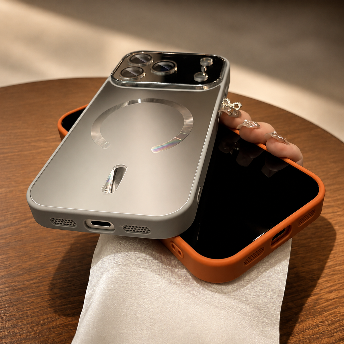 Silver-gray case rests rear-forward across an orange-cased phone in a hand above a wooden table. The lower edge shows charging and speaker openings.