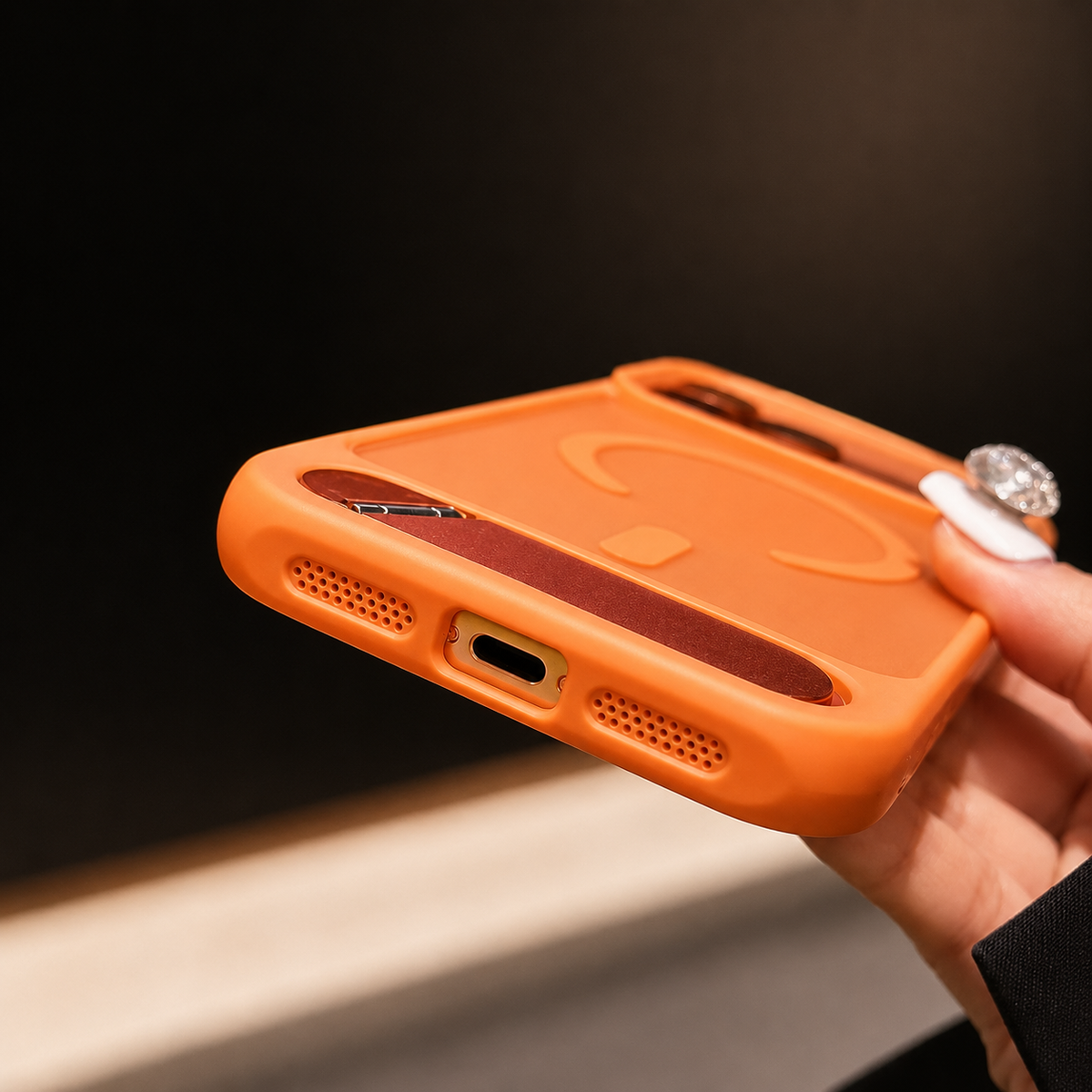 A hand tilts an orange case to show its bottom charging-port opening, speaker holes and rounded frame.
