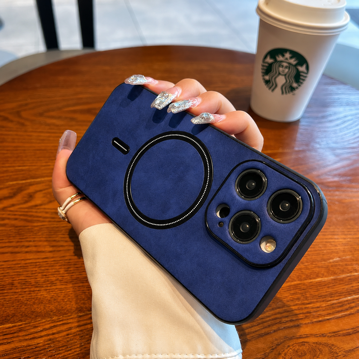 A hand with pale nails and a cream sleeve holds a bright blue stag-emblem case horizontally above a wooden café table. Its dark circular magnetic marking and raised camera panel face the viewer. A white takeaway cup with a green logo sits behind it.