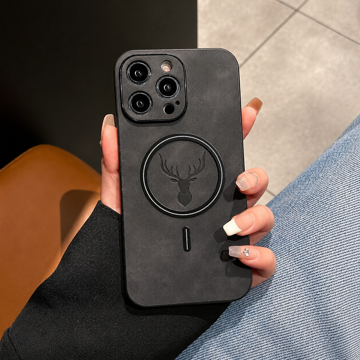 A hand with long pale nails holds a charcoal phone case upright in a café-like setting with a wooden table, tiled floor or upholstered seating behind it. The mottled back has a dark circular ring surrounding an embossed stag head and a short alignment mark below. Matching raised borders surround the camera lenses.