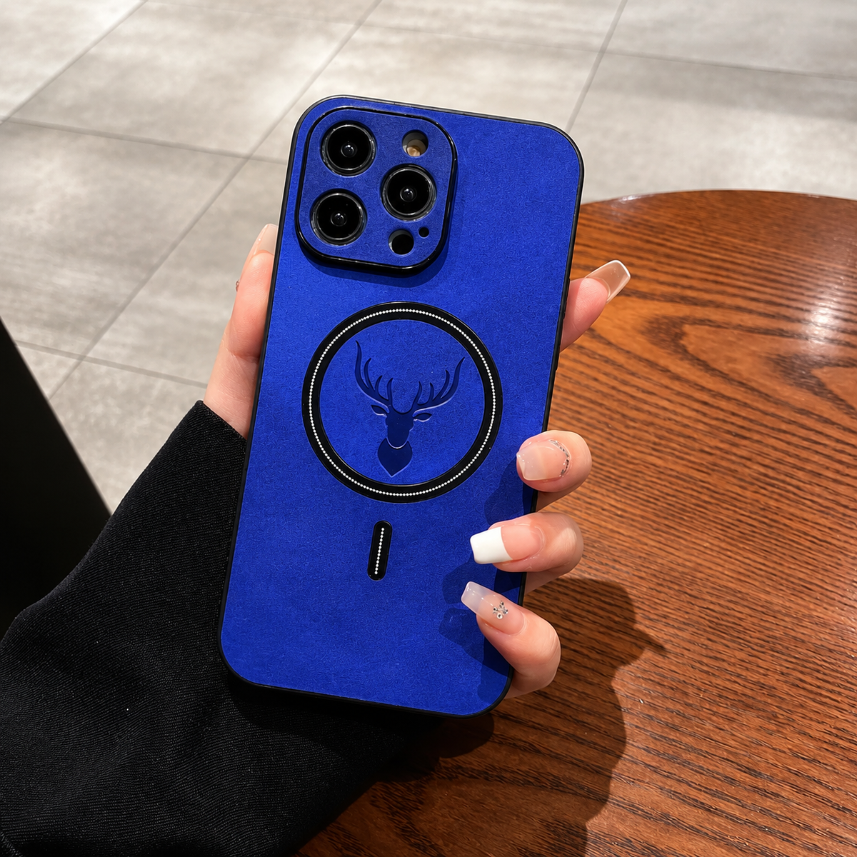 A hand with long pale nails holds a bright blue phone case upright in a café-like setting with a wooden table, tiled floor or upholstered seating behind it. The mottled back has a dark circular ring surrounding an embossed stag head and a short alignment mark below. Matching raised borders surround the camera lenses.