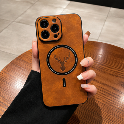 A hand with long pale nails holds an orange and tan phone case upright in a café-like setting with a wooden table, tiled floor or upholstered seating behind it. The mottled back has a dark circular ring surrounding an embossed stag head and a short alignment mark below. Matching raised borders surround the camera lenses.