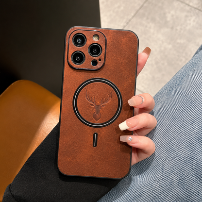 A hand with long pale nails holds a brown phone case upright in a café-like setting with a wooden table, tiled floor or upholstered seating behind it. The mottled back has a dark circular ring surrounding an embossed stag head and a short alignment mark below. Matching raised borders surround the camera lenses.