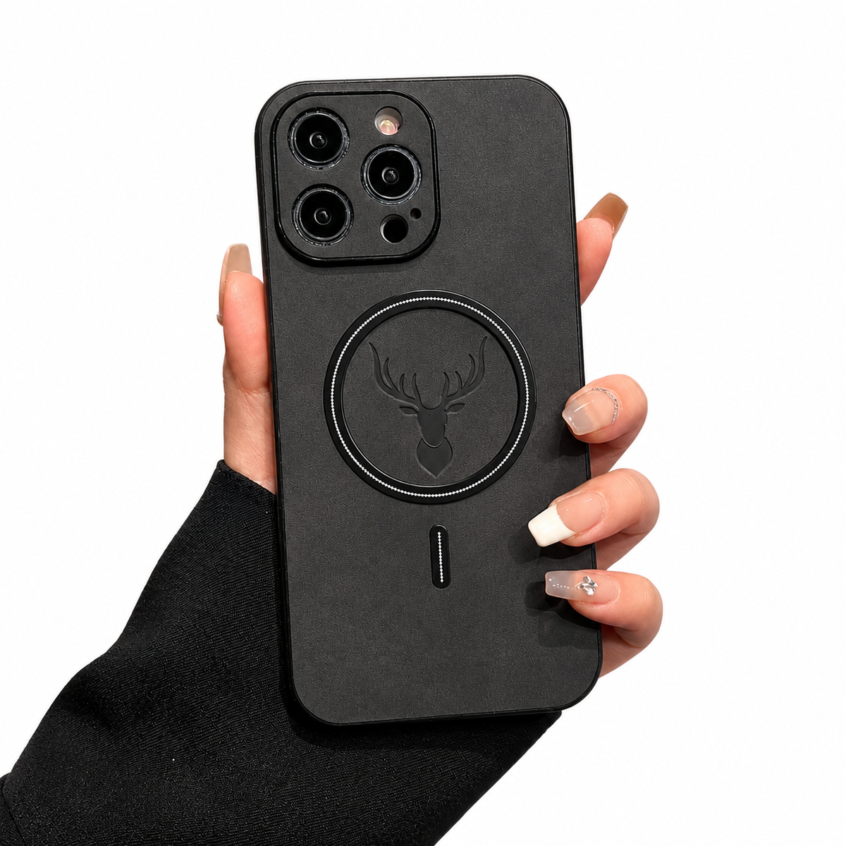 A hand emerging from a black sleeve holds a charcoal phone case upright on white. The soft-looking mottled back has a black circular magnetic ring around an embossed stag head, with a short vertical mark below. Raised dark borders surround the camera lenses at the upper left.