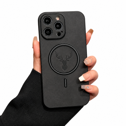 A hand emerging from a black sleeve holds a charcoal phone case upright on white. The soft-looking mottled back has a black circular magnetic ring around an embossed stag head, with a short vertical mark below. Raised dark borders surround the camera lenses at the upper left.