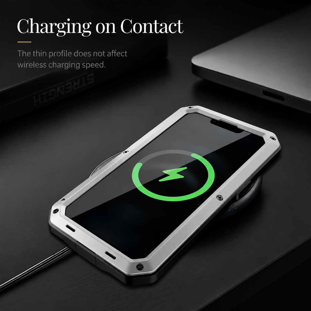 A silver rugged phone lies screen-up on a dark desk with a green charging indicator. Text advertises charging through the case.