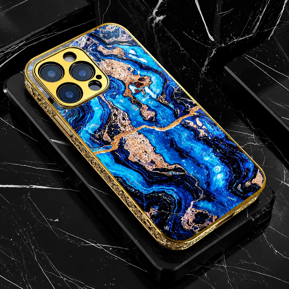 A vivid blue marble-pattern phone case lies on black marble-like blocks. Dark blue swirls and tan-gold patches cover the back, framed by ornate gold-colored edges.