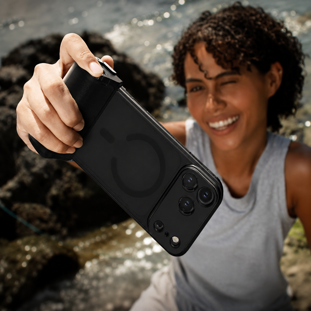 A smiling woman in a gray sleeveless top holds a dark photography case diagonally toward the viewer beside sunlit water and rocks. A camera accessory is attached near the broad camera panel.