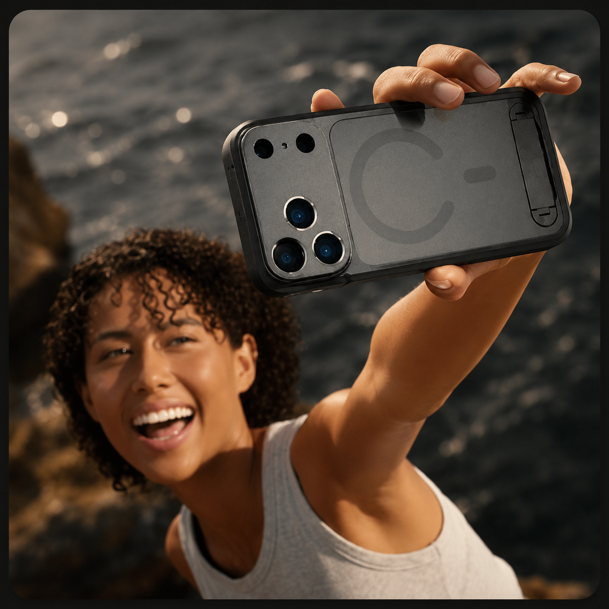 A smiling woman holds a gray photography case overhead for a selfie beside water. The rear circular magnetic marking and broad camera panel face the viewer.