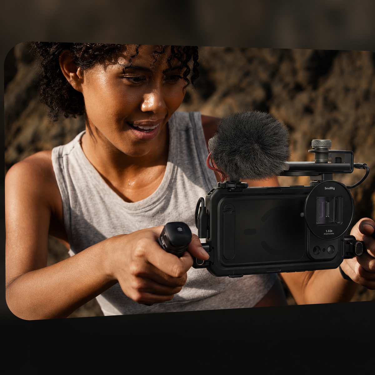 A smiling person in a gray sleeveless top holds a phone horizontally in a dual-handle filming cage beside coastal rocks. A furry microphone and other accessories are mounted above the case. The person looks toward the rig while gripping both handles.