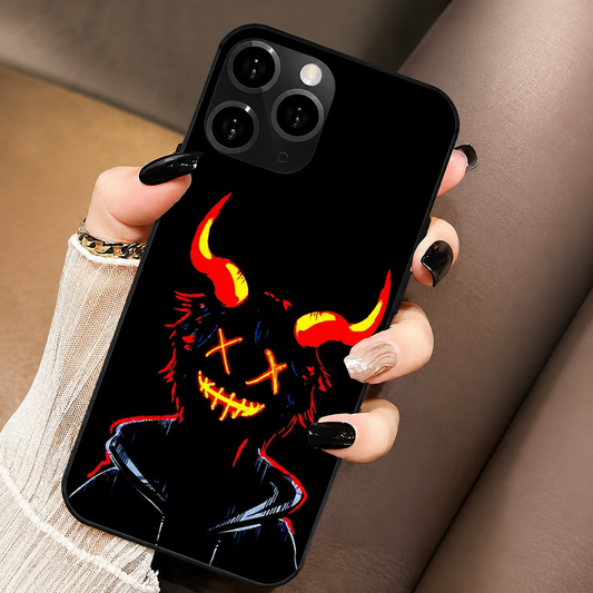 A hand in a white ribbed sleeve holds a black phone with its back facing forward above a brown upholstered seat. The case displays a dark hooded figure with glowing orange cross-shaped eyes, a stitched-looking grin, and red and yellow curved horns. Three camera lenses sit at the upper left. Black polished fingernails and a pale patterned accent nail curl around the phone's edges.