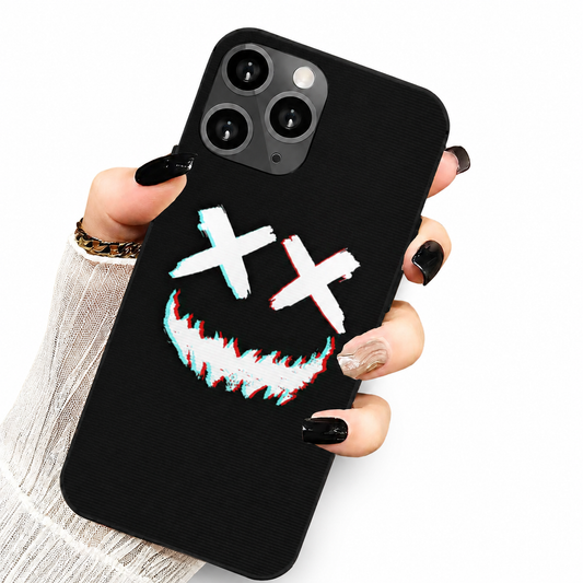 A hand emerging from a white ribbed sleeve holds a black phone upright at a slight diagonal against a white background. The back faces the viewer, with three camera lenses at the upper left and a graphic of two white cross-shaped eyes above a jagged grin edged in red and cyan. The fingernails are mostly glossy black, with one pale patterned nail, and a gold-colored bracelet is visible at the wrist.