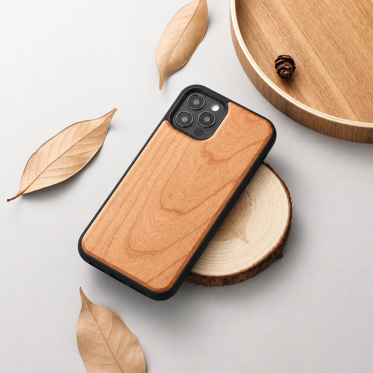 A phone with a honey-brown wood-grain back rests diagonally across a round slice of wood on a light gray tabletop. Three rear camera lenses sit inside a black surround. Dry tan leaves lie to the left and below, while part of a wooden tray holding a small cone appears at the upper right.