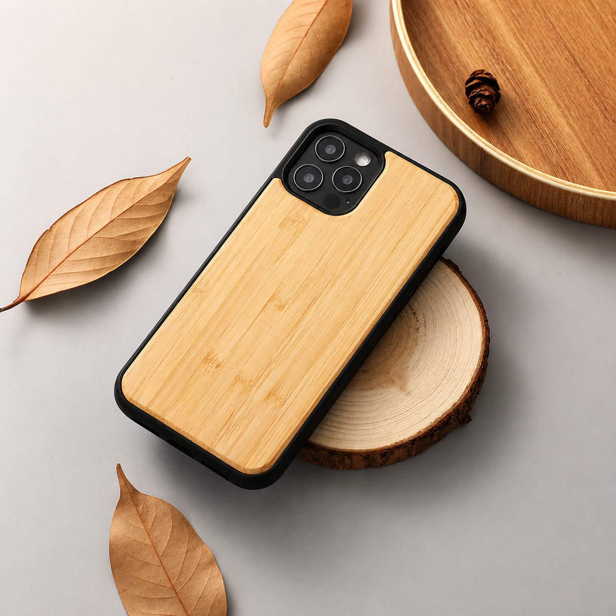 A phone with a pale golden wood-grain back lies diagonally across a round wooden slice on a light gray surface. A black border frames the case and its three-lens camera opening. Dry tan leaves surround the arrangement, with part of a wooden tray and a small cone at the upper right.