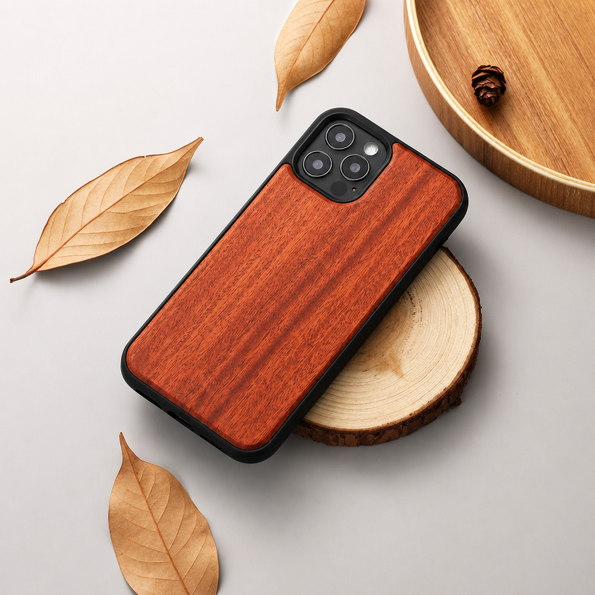 A phone with a reddish-orange wood-grain back lies diagonally over a round wood slice. Its black edges and three-lens camera surround contrast with the streaked panel. Dry tan leaves lie on the light gray tabletop, and a curved wooden tray with a small cone enters from the upper right.