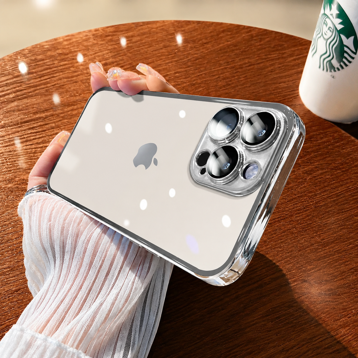 Hand holds a clear silver-colored case horizontally above a wooden table. The pale phone and logo show through the back beneath bright reflections.