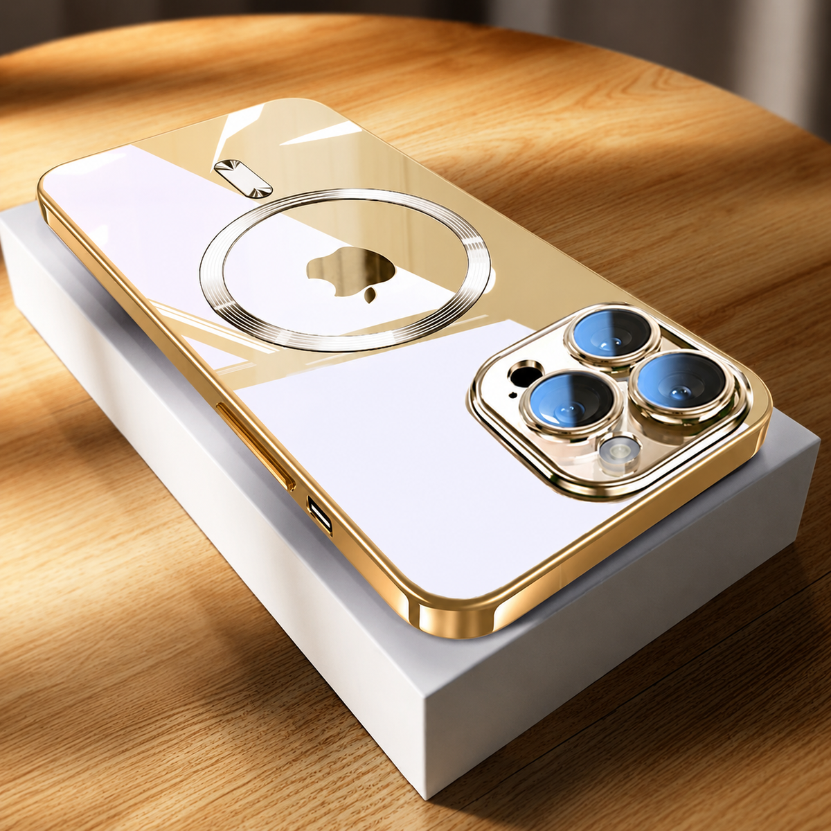 A gold-edged phone rests diagonally on a white rectangular box on a sunlit wooden tabletop. Its pale reflective back shows a circular gold-toned ring around the phone logo and a short bar above. Three camera lenses with blue reflections sit within a gold-colored surround at the nearest end.