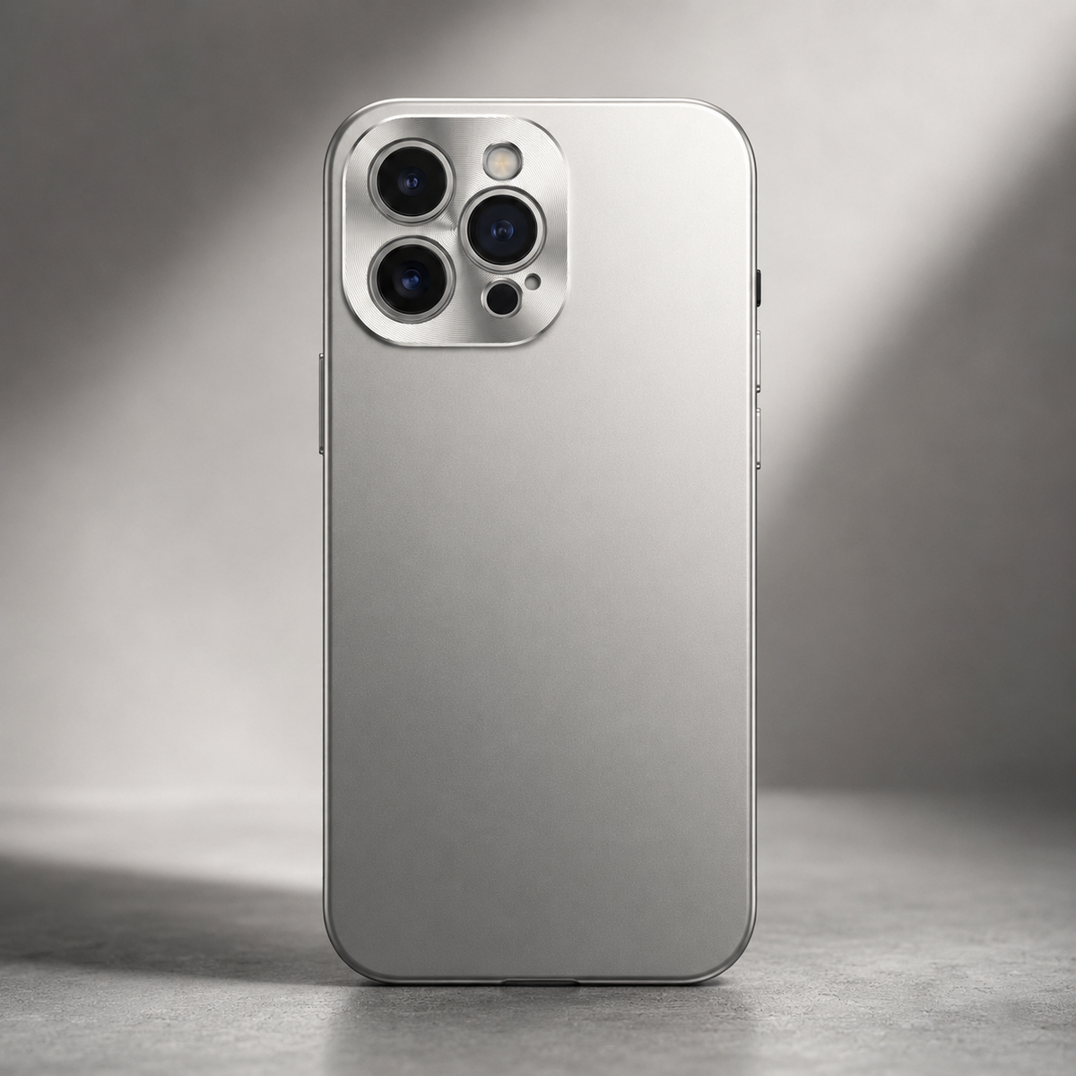 A silver case stands upright on a gray surface in soft light. Its smooth matte back and rounded-square three-lens camera surround face the viewer.