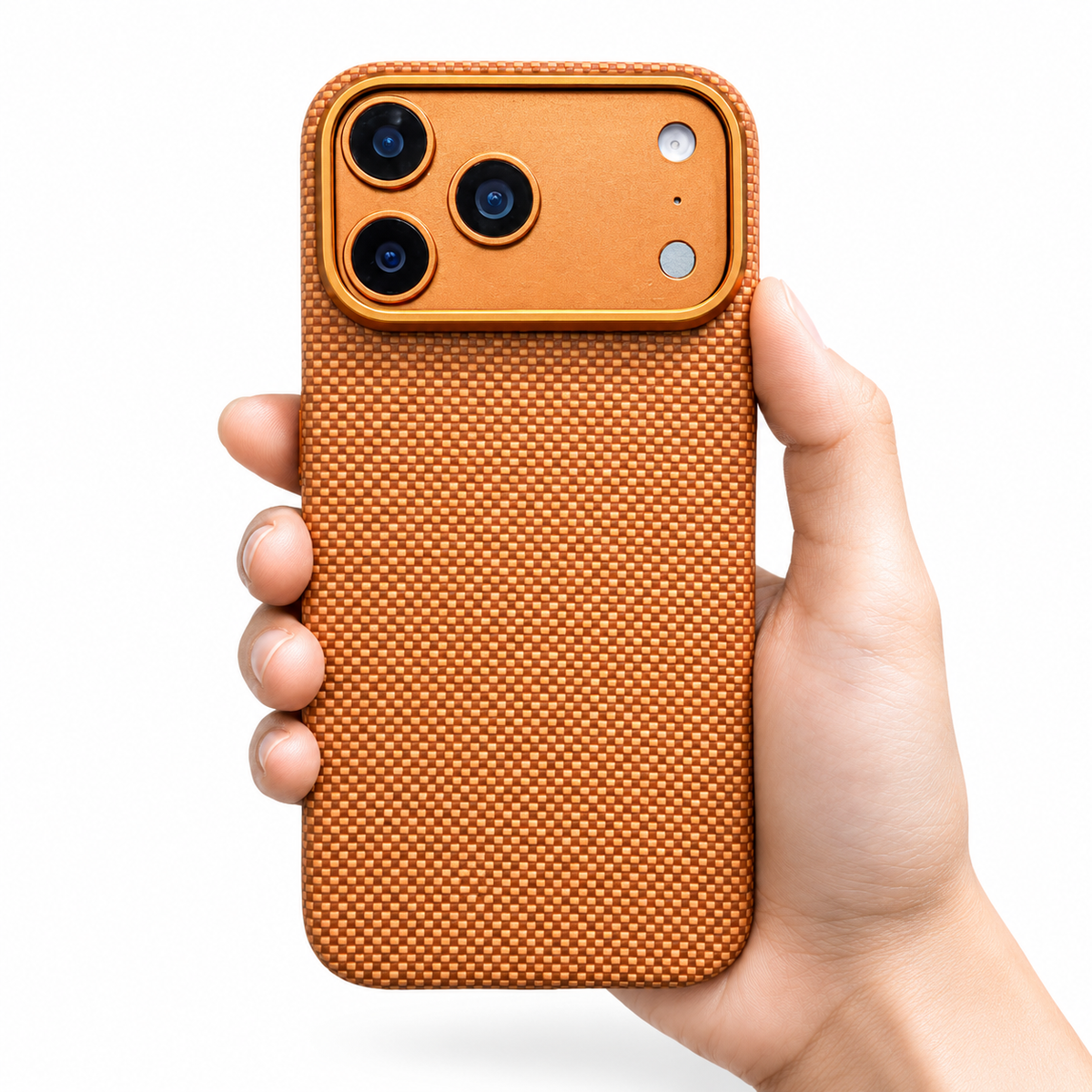 A hand holds an orange-gold fine-weave phone case upright on white. A broad matching camera panel with three black lenses sits above the densely interlaced back pattern. A thin gold-orange rim outlines the rounded corners and camera panel, and the hand supports the lower edge.