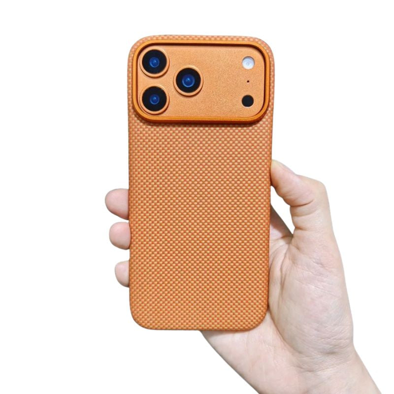 A hand holds a muted orange fine-weave phone case upright on white. A broad matching camera panel with three black lenses sits above the densely interlaced back pattern. A thin gold-orange rim outlines the rounded corners and camera panel, and the hand supports the lower edge.